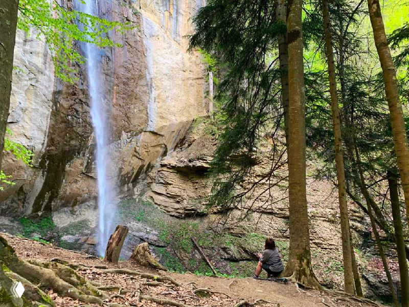 Посетитель сидит на камне у подножия водопада, вокруг влажные камни и зелень, ощущение масштабов стены воды.