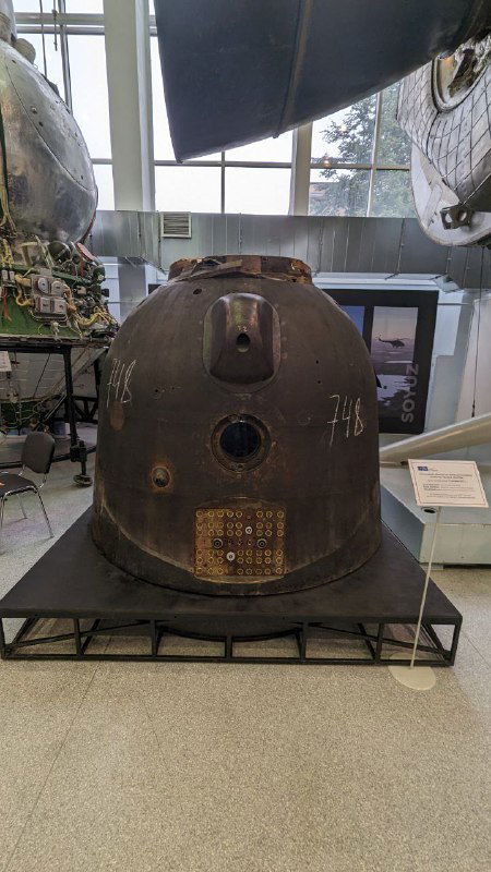 Спускаемый аппарат в музее Роскосмоса: металлическая обшивка, следы шаровидного корпуса и табличка, экспонат, связанный с возвращением экипажей.