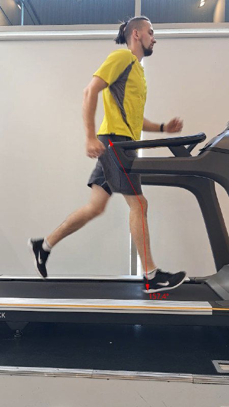 Боковой снимок человека в жёлтой футболке, бегущего на беговой дорожке в зале: видна постановка стопы, шаг и работа кроссовок при тестировании техники.