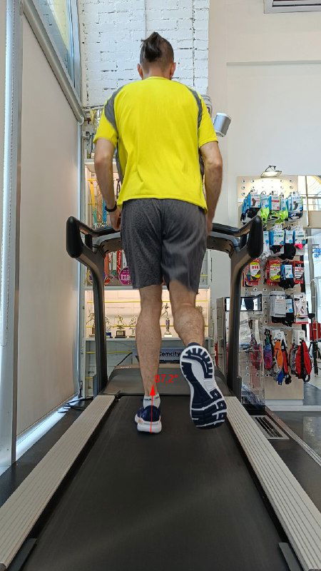 Сзади: бегун в жёлтой футболке и серых шортах на дорожке в магазине, видно пятку и подошву кроссовок на фоне торговых стеллажей.