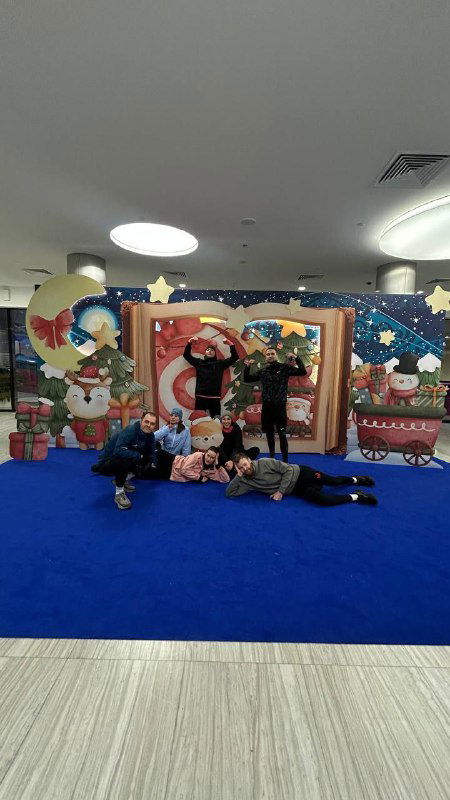 Праздничная фотозона с Дедом Морозом и украшениями в торговом центре, рядом люди и участники мероприятия на синем полу.