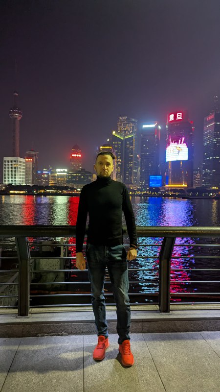 Человек у перил на набережной вечером на фоне подсвеченных высотных зданий и рекламных вывесок, городской портрет в сумерках с рекой позади.