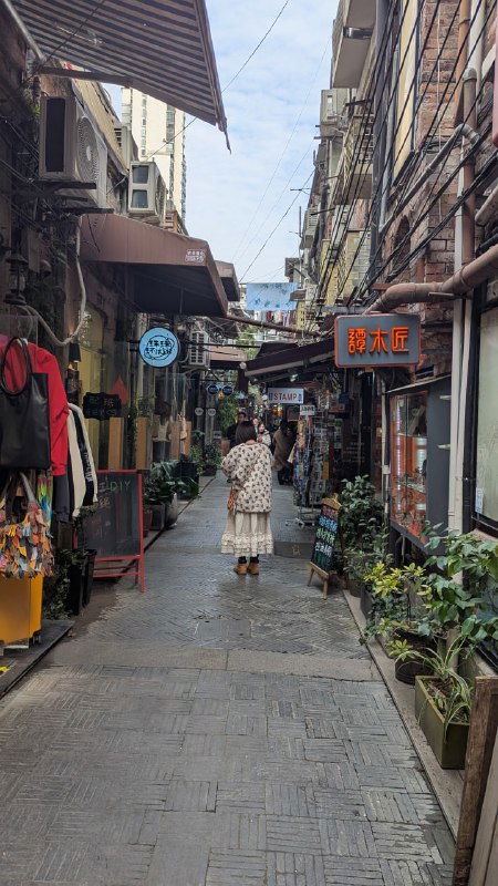 Узкая пешеходная улочка старого района с низкими зданиями и витринами, прохожий идёт по центру — повседневная сцена городской жизни и небольших лавок.