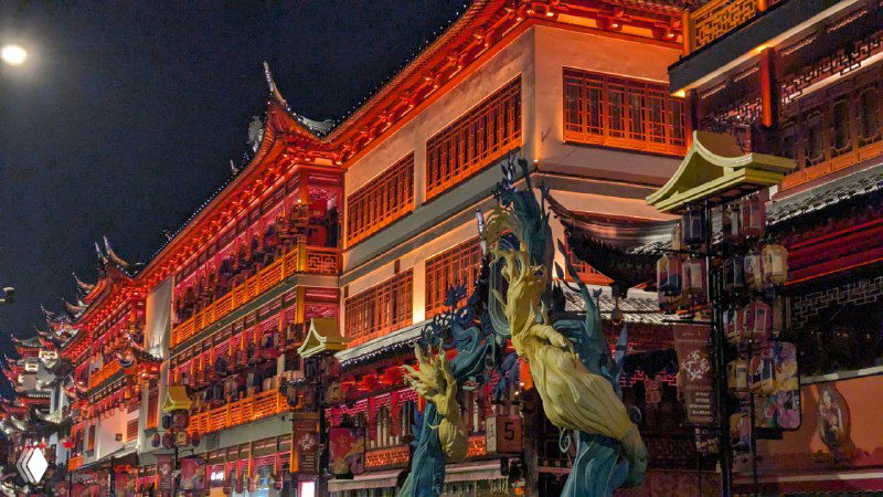 Ярко подсвеченное традиционное азиатское здание с архитектурными деталями и статуями, вечерний снимок храма или исторической части города с насыщенной подсветкой.