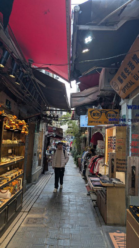 Узкий торговый переулок с множеством прилавков, вывесок и навесов, люди прогуливаются мимо мелких лавок — уличный рынок и коммерческая жизнь района.