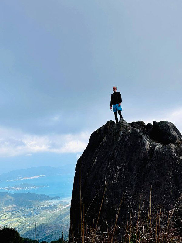 Автор стоит на вершине утеса или утёса в горах, позади открывается панорама побережья и облачное небо — ощущение свободы и высоты.
