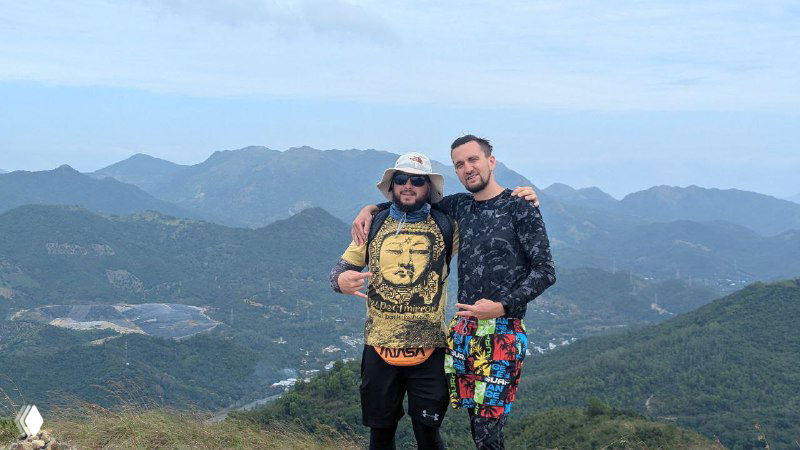 Двое мужчин на фоне горной панорамы — один держит тематическую футболку, оба улыбаются; снимок с похода в горы, дружеская атмосфера.