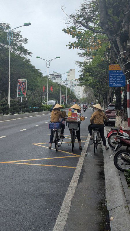 Улица города с вьетнамскими уличными торгами: велосипедисты и продавцы в традиционных шляпах, городской ритм и уличная жизнь в кадре.