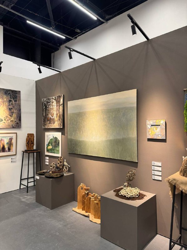 Стенд галереи с большими спокойными пейзажными полотнами и керамическими или деревянными объектами на подиуме внизу.