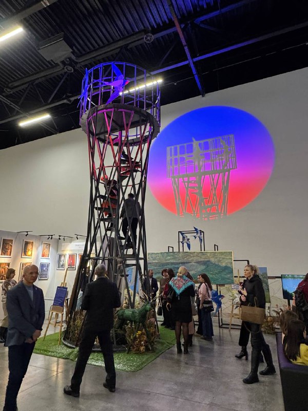 Пространство выставки с крупной инсталляцией в центре и посетителями, которые обходят экспозицию под высокими потолками павильона.