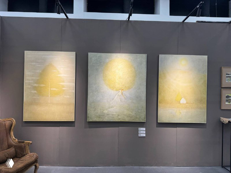 Три крупные мягкие по тону абстрактные картины висят рядом на нейтральной стене; внизу небольшой стул и элементы оформления стенда.