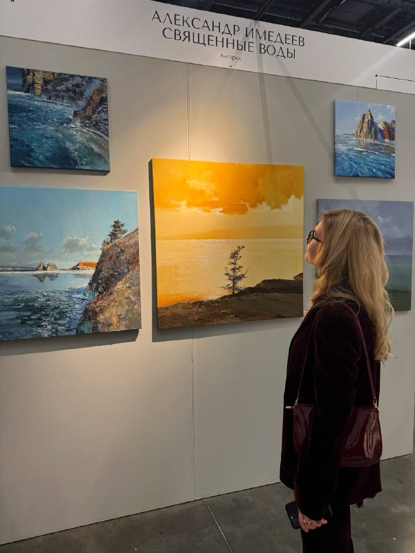 Посетительница стоит лицом к стенду с несколькими пейзажными картинами, одна из которых подсвечена тёплым светом — кадр передаёт процесс созерцания.