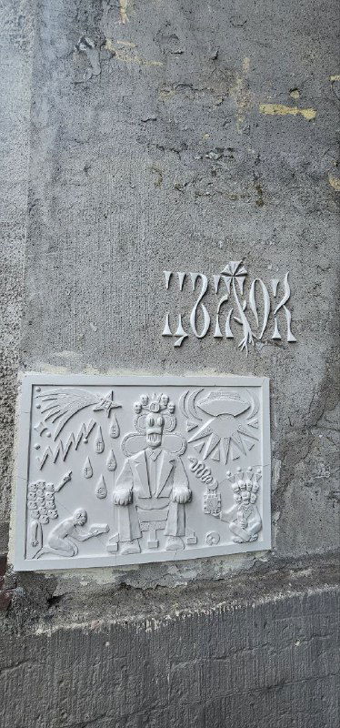 Узкий вертикальный снимок рельефного табло и декоративной плитки на фасаде; видно небольшое стилизованное слово и сюжетную плиту внизу.