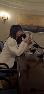 Женщина в зале конференции делает снимок на смартфон во время сессии, видно столы, микрофоны и часть аудитории, рабочая атмосфера.