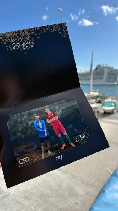 Буклет или открытка с фотографиями и сувенирной вставкой, лежащая на фоне морского порта и пришвартованного лайнера в лучах солнца.