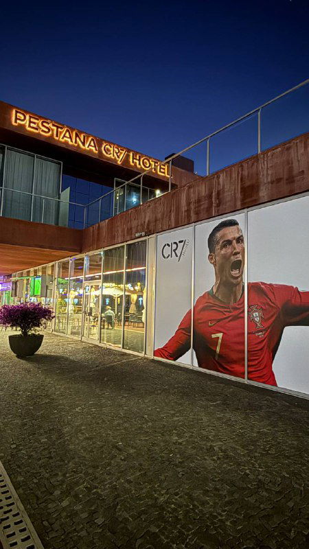 Вечерний фасад отеля или ресторанного комплекса с большими баннерами и портретными изображениями знаменитости на наружной стене у набережной.