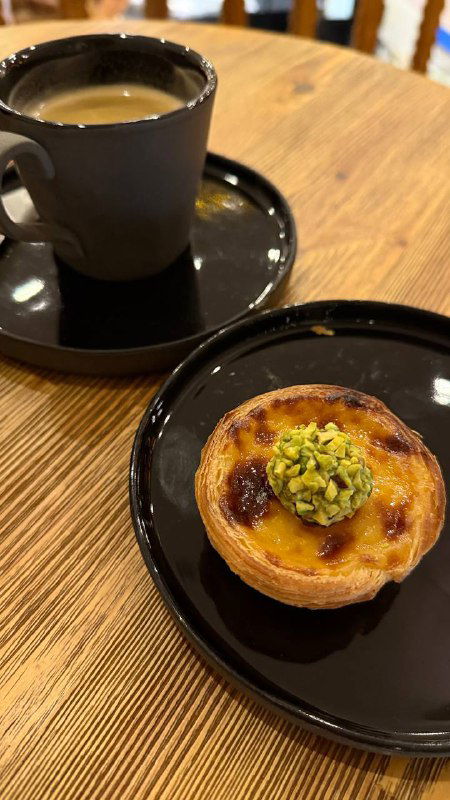 Чёрная чашка с кофе и маленькая тарелка с пастейш, сверху украшение из фисташек — крупный план десерта в уютном кафе.