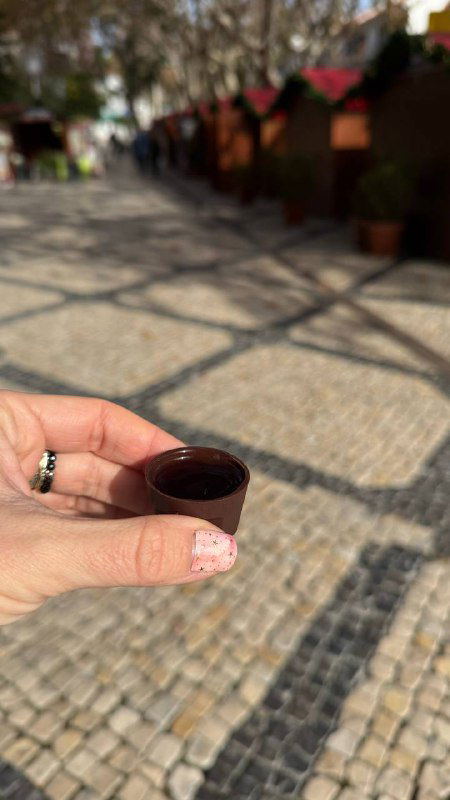 Рука держит маленькую шоколадную чашечку с ликёром жижинью на мощёной площади; размытый городской фон и солнечный день.