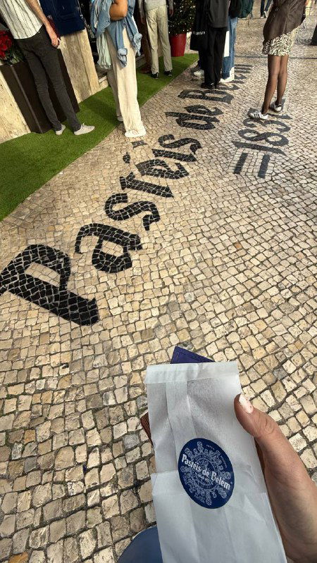 Мозаичный мощёный тротуар с надписью 'Pastéis de Belém' и рука держит бумажный пакет с логотипом кондитерской — вид у входа.