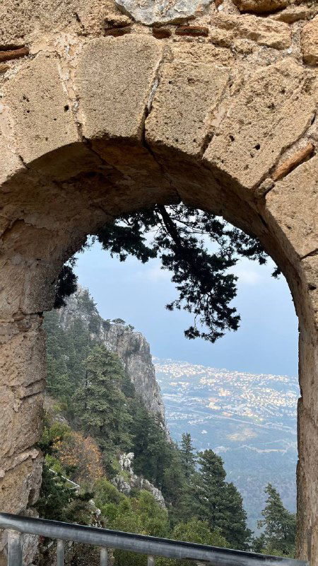 Вид через каменную арку на морской пейзаж и прибрежные склоны — ракурс с высоты, показывающий природу и город вдали.