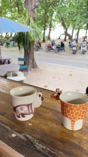 Две керамические чашки на столе с видом на песчаный пляж и набережную: детали сервировки и уличная сцена пляжа Най Янг с прохожими и зонтиками.