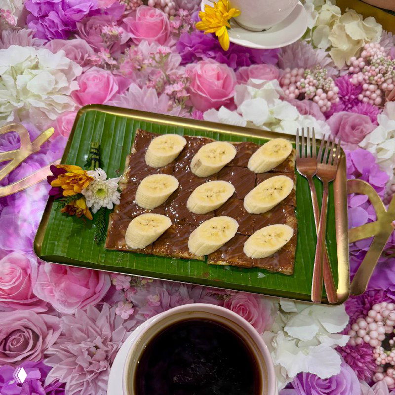 Десерт на зелёной банановой пластине и чашка кофе на фоне розово-лиловых искусственных цветов в стиле кафе Kor Ra Kang — яркая фотозона.