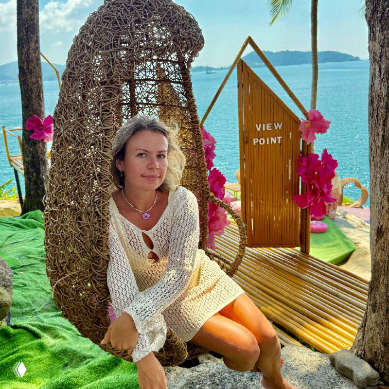 Туристка сидит в плетёном коконном кресле у фотолокации с табличкой "VIEW POINT" на фоне моря — типичная инста-позировка.