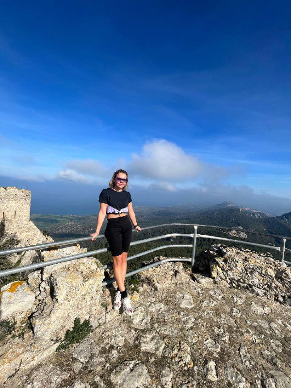 Я у руин замка на вершине холма, вид на зелёные склоны и море — фотография туристки на фоне средиземноморского пейзажа и крепостной стены.