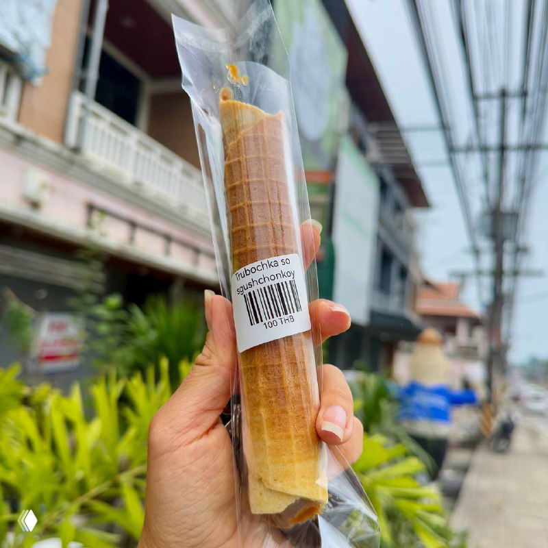 Рука держит упакованную трубочку с начинкой на улице Самуи: пластиковая упаковка, штрих-код и городская улица на фоне.