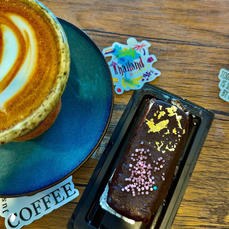 Флет-вайт в синей чашке рядом с шоколадным сырком, украшенным съедобным золотом и посыпкой, на деревянном столе в кафе.