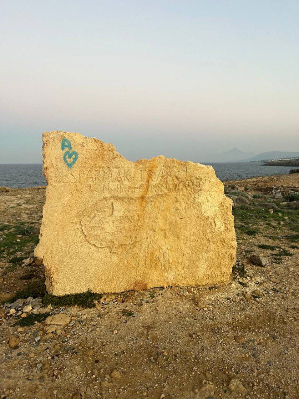 Кусок каменной плиты с бирюзовым граффити у береговой линии, на заднем плане море и линия горизонта — небольшой береговой артефакт.