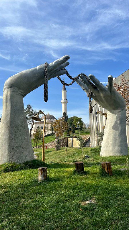 Большая скульптура в виде рук, сцена инсталляции на лужайке острова — арт-объект на тему свободы и независимости, яркое небо на фоне.