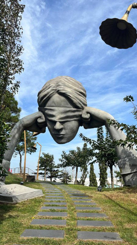 Скульптурная голова человека с закрытыми глазами в парковой зоне острова, дорожки и деревья вокруг, арт-инсталляция в натуральном окружении.