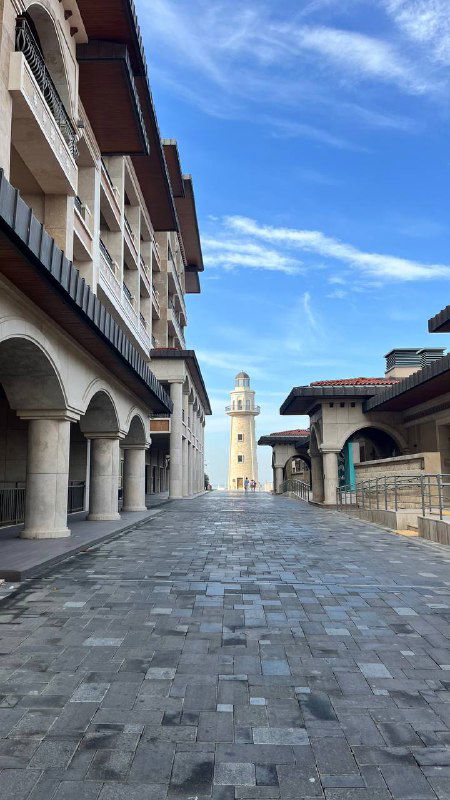 Пустая просторная улица островного комплекса с зданиями и башней на конце — архитектурные элементы отеля/музейной зоны при ясном небе.