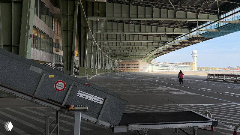 Пандус и часть летного поля Темпельхофа под навесом ангаров, на фоне видна взлётно-посадочная зона и небольшая активность на площадке.