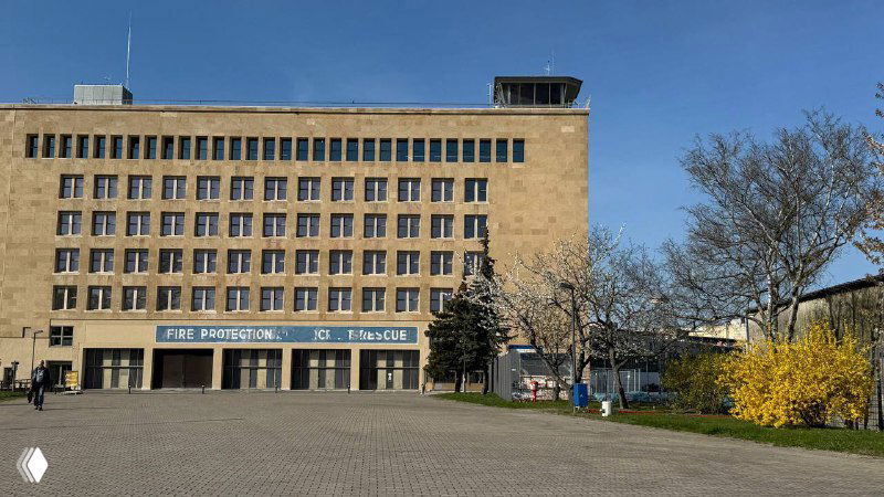 Вид на фасад административного здания Темпельхофа с прилегающей площадью и деревьями — монументальная городская перспектива.