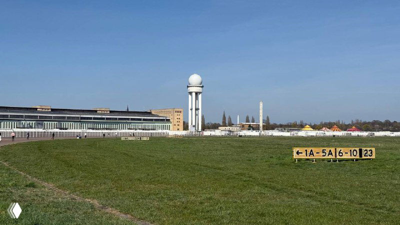 Большая зелёная полоса бывшего летного поля Темпельхофа с наблюдательной башней и промышленным горизонтом Берлина на заднем плане.