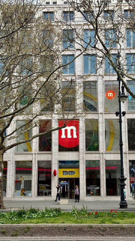Внешний вид трёхэтажного магазина M&M's в Берлине с большим красным логотипом M и витриной на улицу через деревья и тротуар.