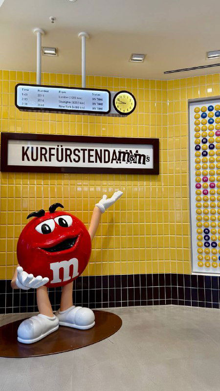 Интерьер M&M's: жёлтая плиточная стена, вывеска Kurfürstendamm и ростовая фигура красного персонажа M&M для фото у посетителей.