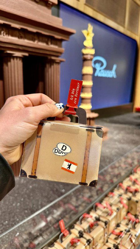 Рука держит небольшой сувенир в виде чемоданчика с шоколадной упаковкой и ценником в магазине Rausch, кадр крупным планом на фоне витрин.