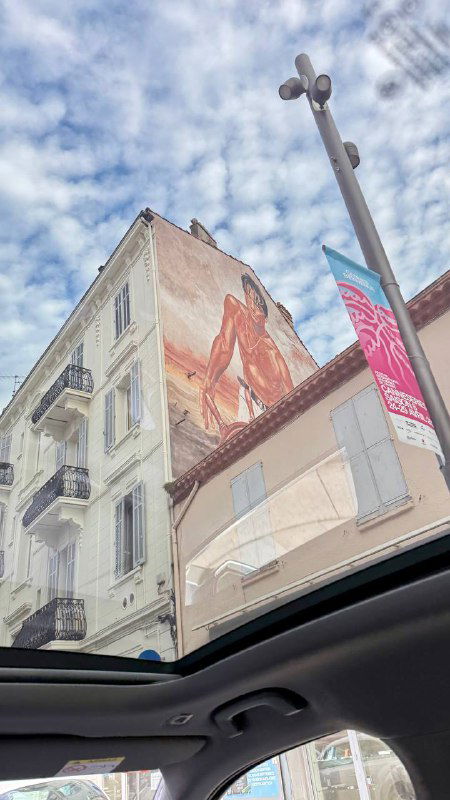 Вид из машины на фасад здания с большой настенной росписью и небом в отражении крыши — урбанистическая деталь Канн.