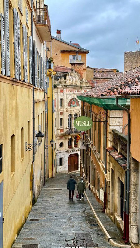 Узкая мощёная улица Мужена с высокими домами по бокам, вывеской музея и несколькими прогуливающимися людьми вдалеке.