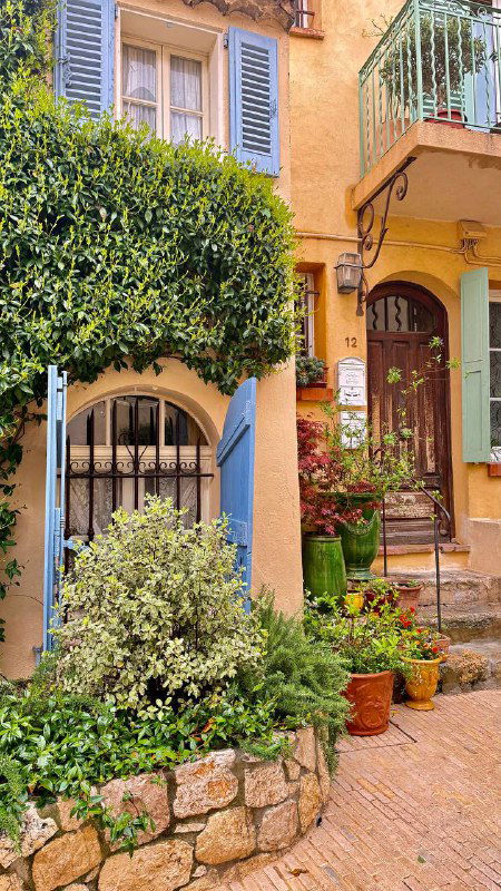 Яркий уголок с домом в тёплых оттенках, ставнями на окнах и множеством цветочных горшков у входа — уютный фасад в Провансе.