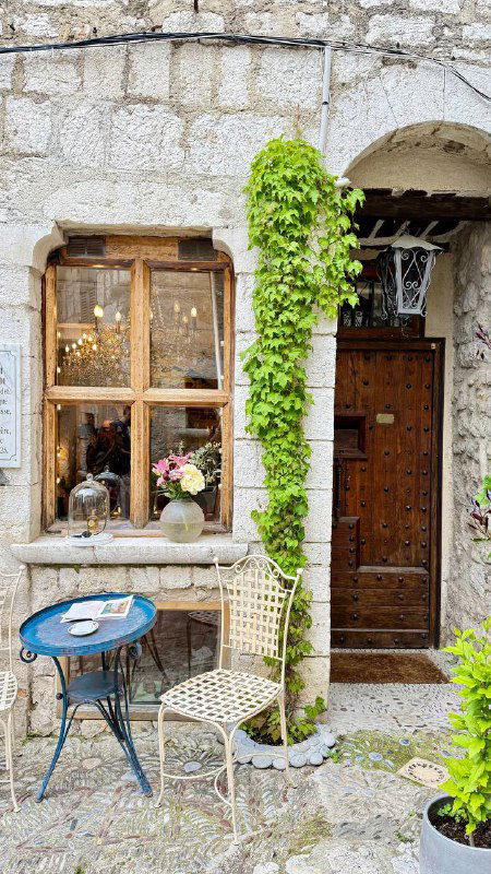 Уютный уголок в старинном доме Сен‑Поль‑де‑Ванс: столик на улице, стул и окно с цветами, атмосфера деревни‑арт.