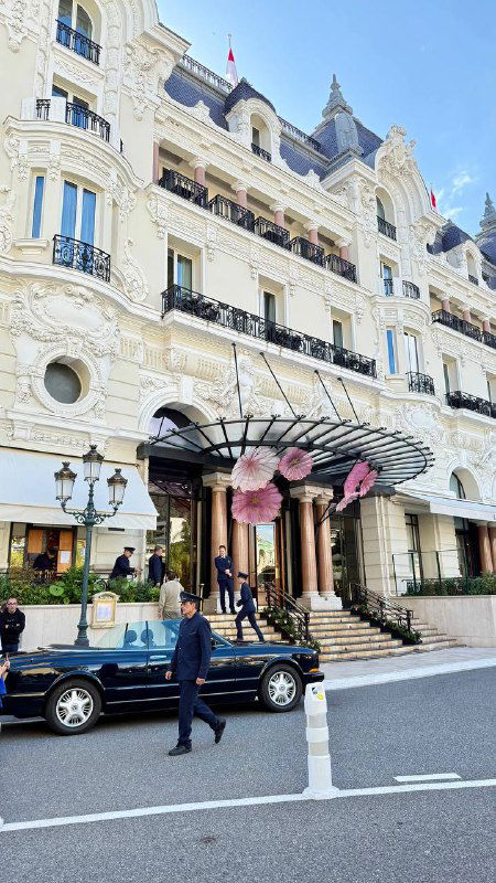 Фасад казино в Монте‑Карло: роскошное здание с колоннами, вывесками и проезжающими автомобилями на переднем плане.