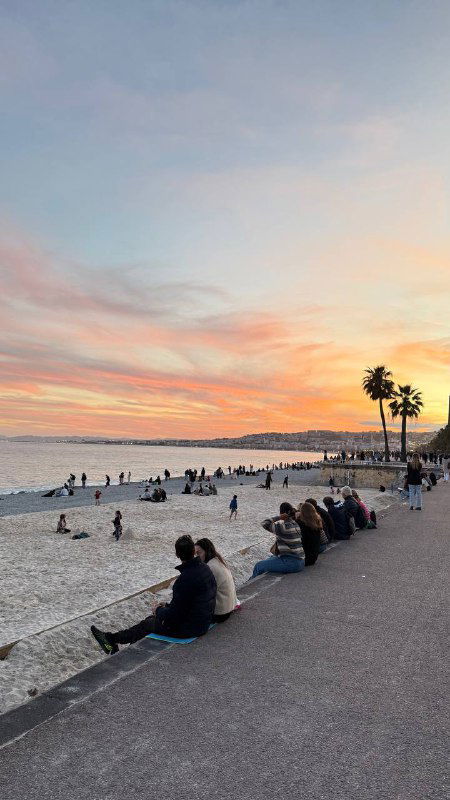 Закат на набережной Ниццы: люди сидят у моря, небо в розово‑оранжевых тонах и силуэты пальм на горизонте.
