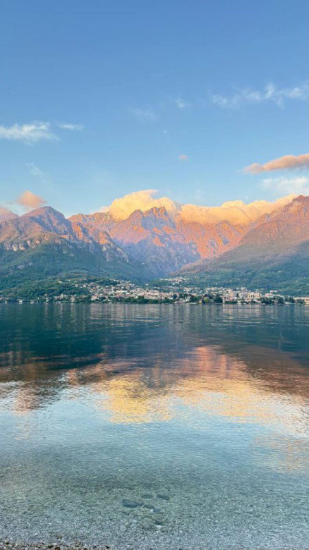 Спокойная водная гладь озера Комо отражает горы и нежные облака при тёплом свете — гармоничный пейзаж с отражением деревень на берегу.