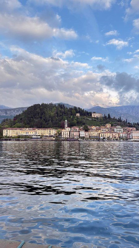 Прибрежный городок на противоположном берегу озера: набережная, здания и спокойная водная поверхность под светлым облачным небом.