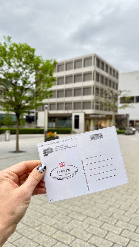 Рука держит открытку с поставленным сувенирным штампом Лихтенштейна на фоне городских домов и офисного здания.