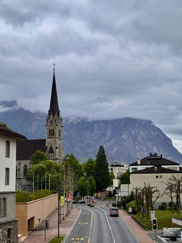 Городская улица Лихтенштейна: вид на церковь со шпилем, пустую дорогу и горы на фоне под облачным небом в пасмурный день.